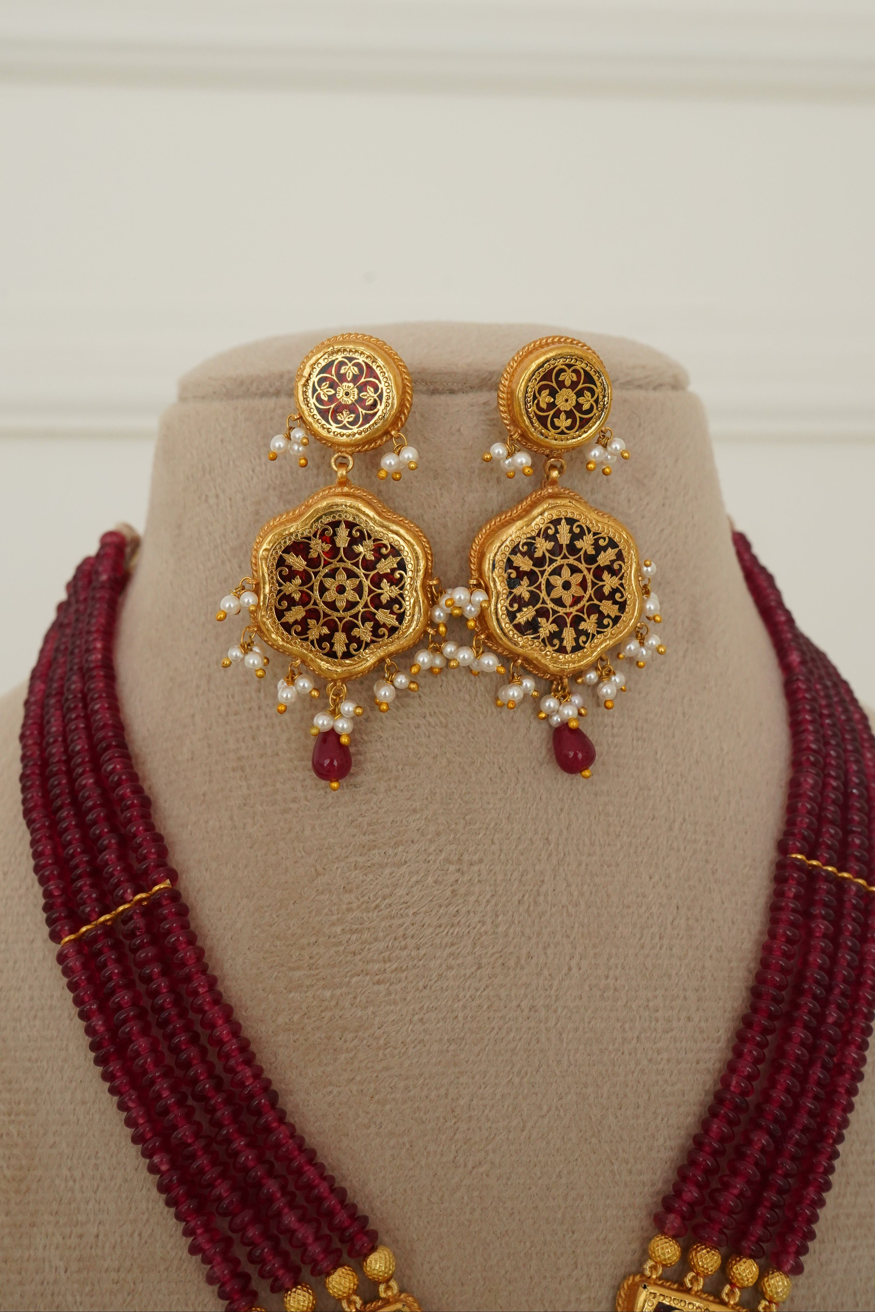 Traditional Indian jewelry set featuring a deep maroon beaded necklace, matching earrings, and a gold-plated necklace with intricate black and gold floral engravings and small white pearls, displayed on a beige mannequin bust.