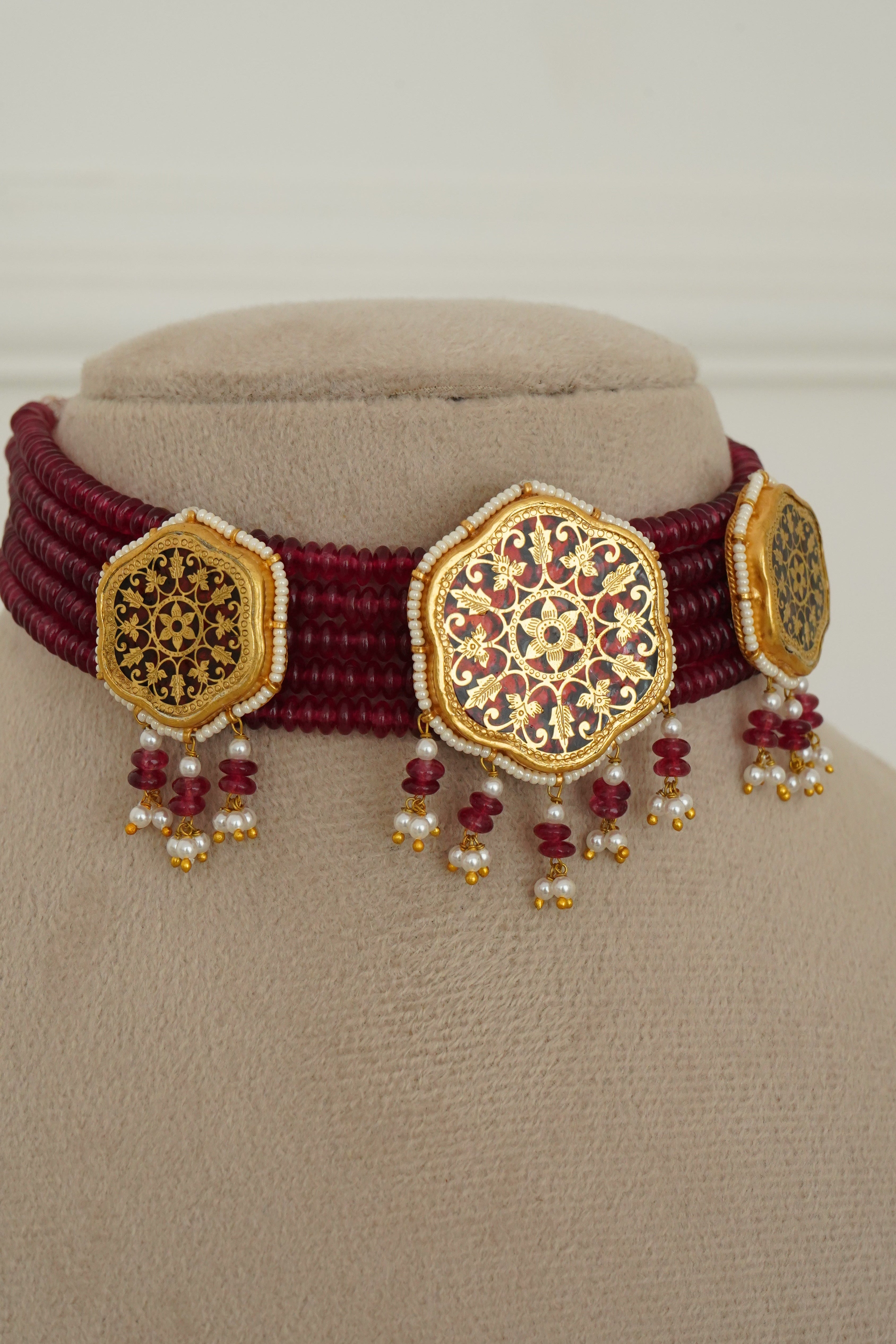 A close-up of a traditional Indian jewelry piece featuring a deep maroon woven band with three ornate gold-plated hexagonal plates decorated with intricate red and gold floral patterns, embellished with small red and white bead tassels, displayed on a beige jewelry stand.