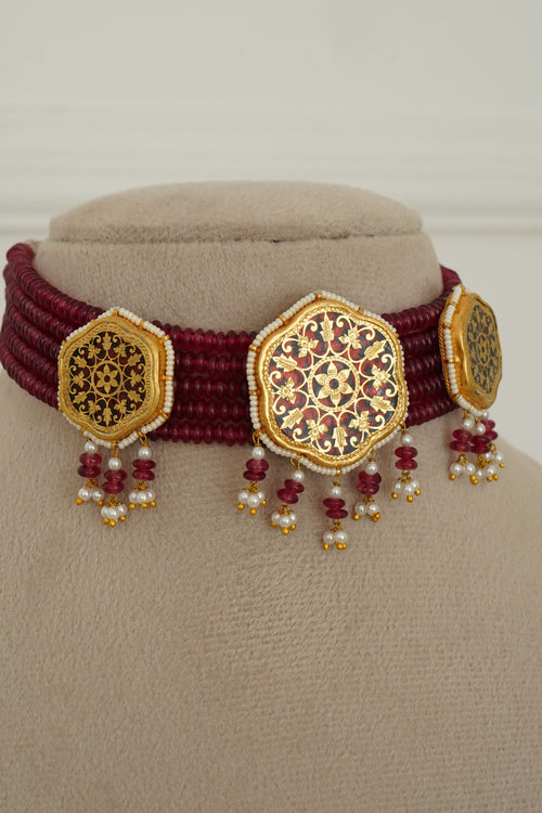 A close-up of a traditional Indian jewelry piece featuring a deep maroon woven band with three ornate gold-plated hexagonal plates decorated with intricate red and gold floral patterns, embellished with small red and white bead tassels, displayed on a beige jewelry stand.