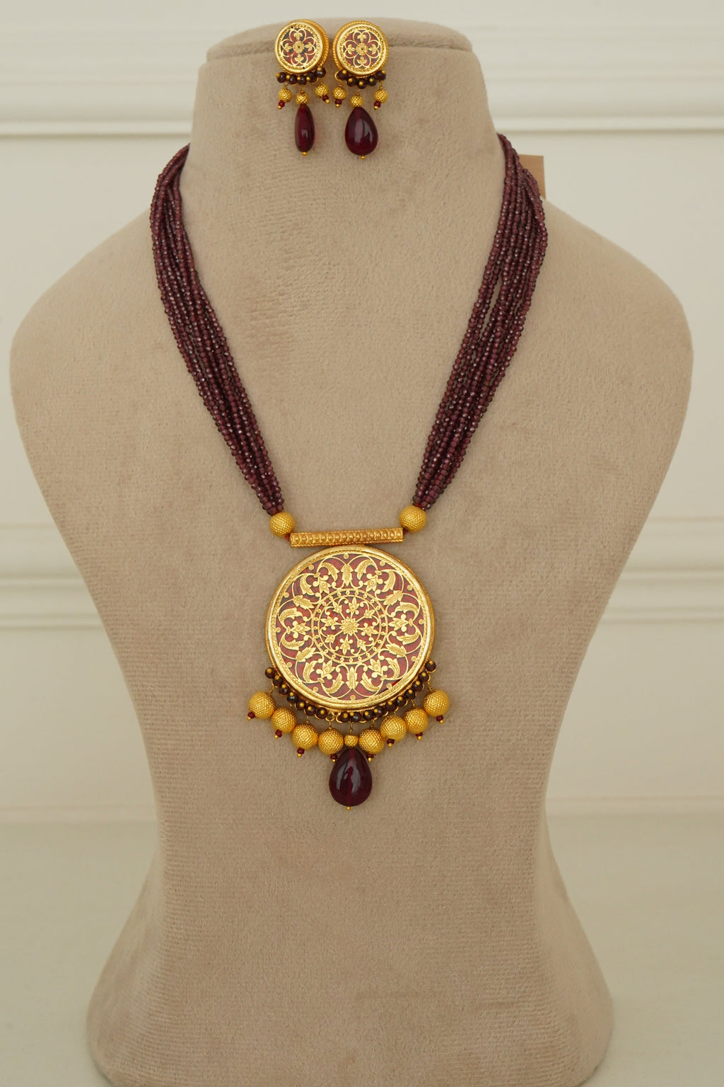 A traditional Indian statement necklace and matching stud earrings displayed on a beige velvet jewelry bust. The necklace features a large, ornate gold-toned circular pendant with intricate filigree work, accented by small gold beads and dark purple teardrop crystals.