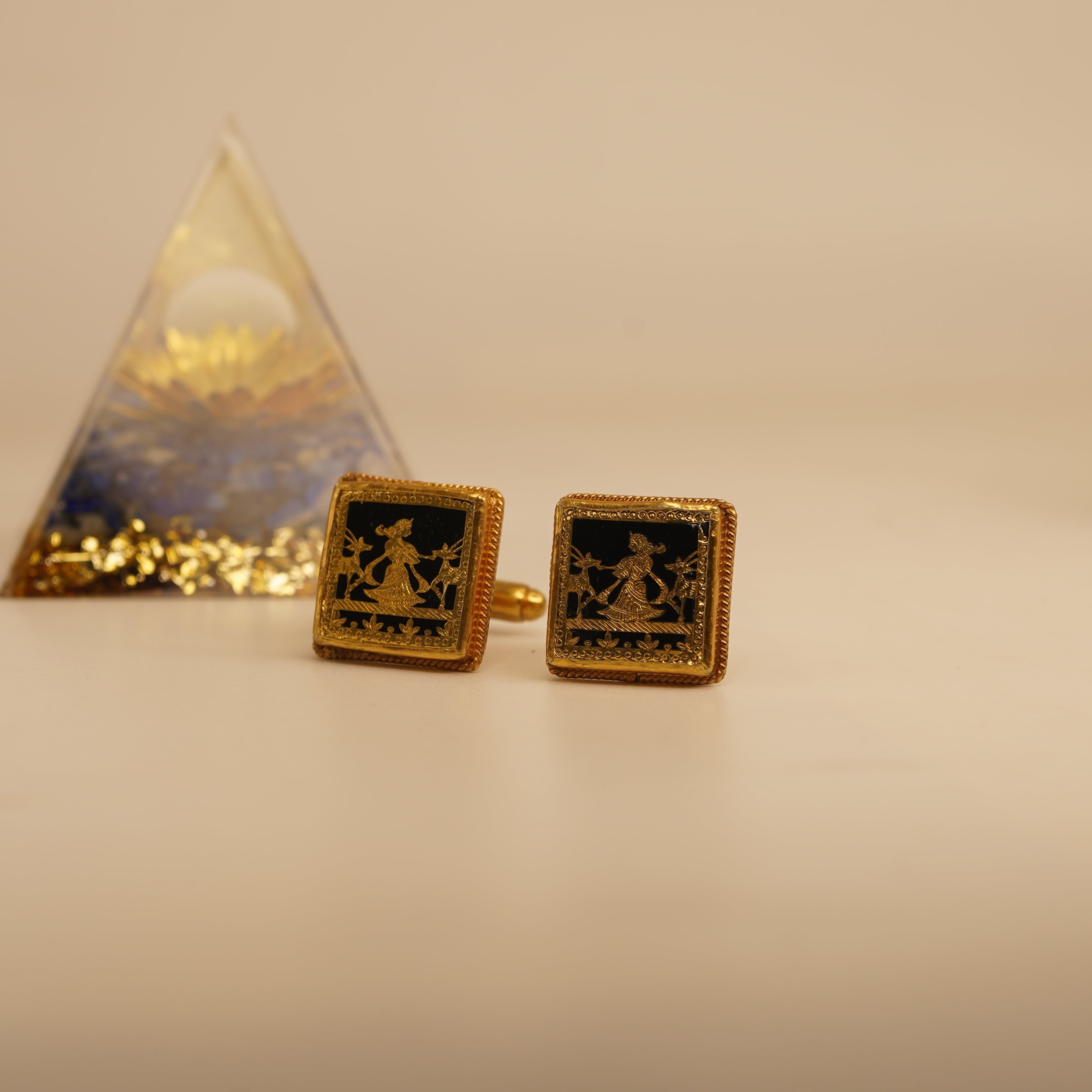 Gold cufflinks with a pyramid-shaped object in the background on a light surface.